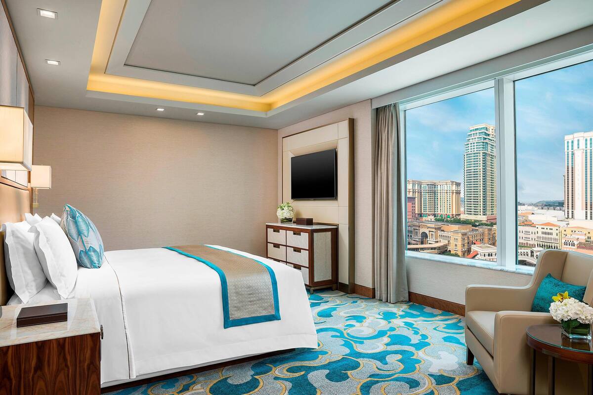 A hotel room with a bed, a desk, a chair, a TV, and a large window with a city view.