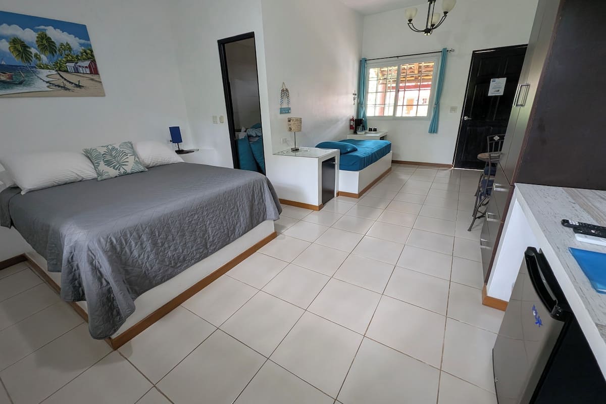Habitación de hotel con cama, escritorio, silla, una mesita, ventana con cortinas y un cuadro en la pared.