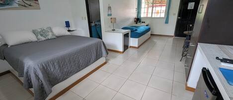 Habitación de hotel con cama, escritorio, silla, una mesita, ventana con cortinas y un cuadro en la pared.