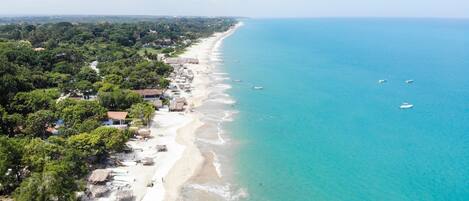 An aerial view of a beach with clear turquoise waters, a sandy shore, and scattered buildings along the coastline.