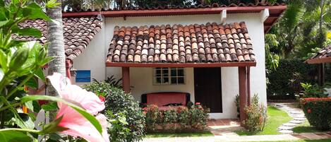 Un pequeño edificio con techo de tejas, rodeado de abundante vegetación y un jardín cuidado.