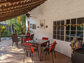 An outdoor patio with a glass table and wicker chairs, a ceiling fan, and a window with a view of parked cars. - Villa Botero By Casa Mojito B&B (Rio Hato)