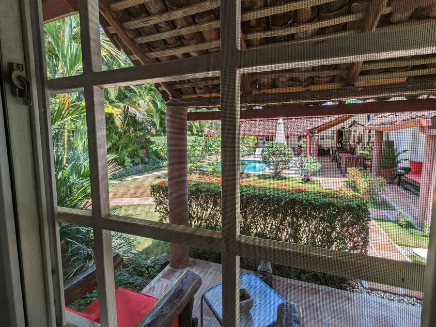 A view through a window of a covered outdoor area with a pool, seating, and greenery.