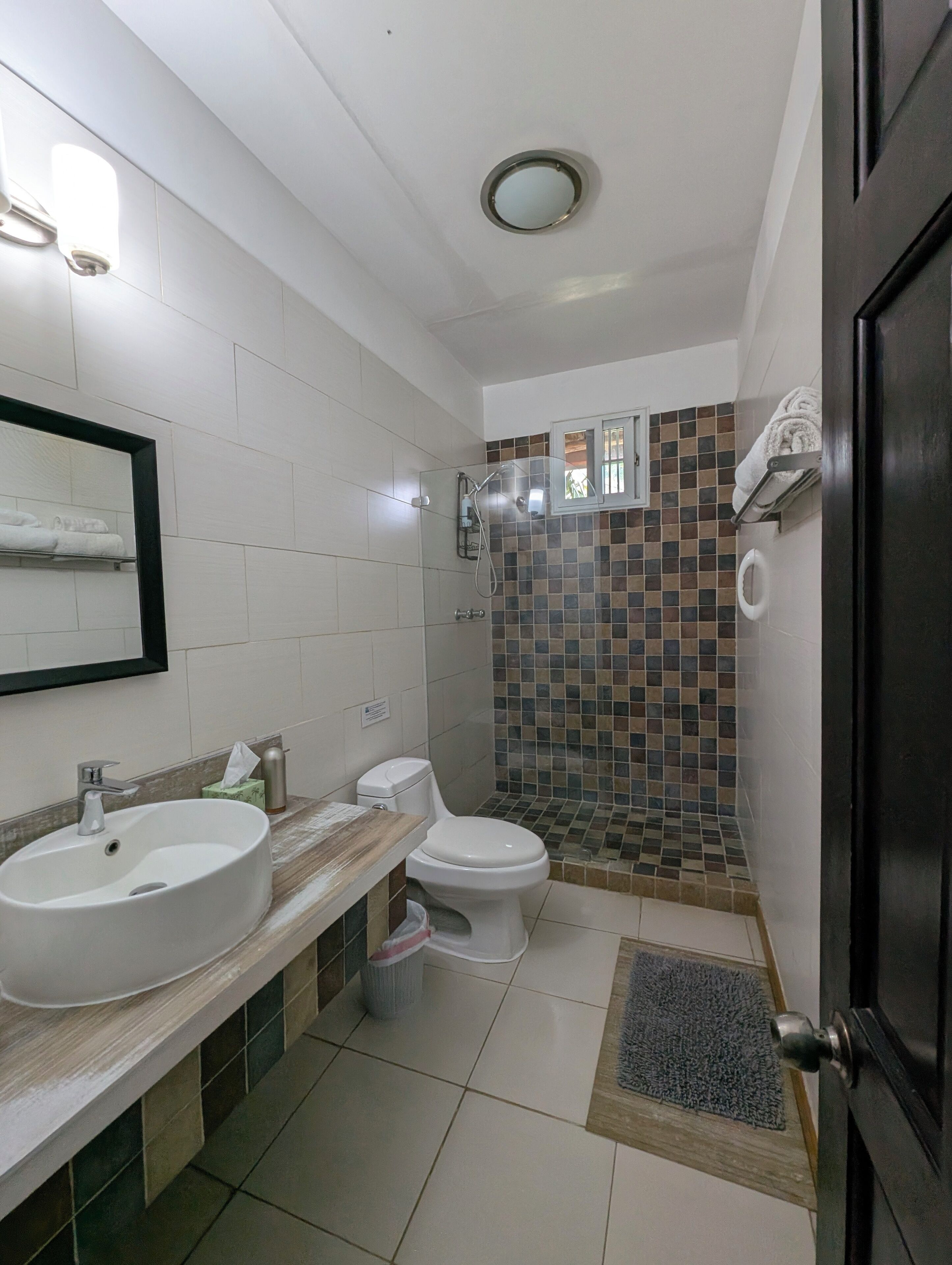 Un baño con lavamanos, inodoro y ducha blancos. Hay un espejo sobre el lavamanos y un perchero encima del inodoro.