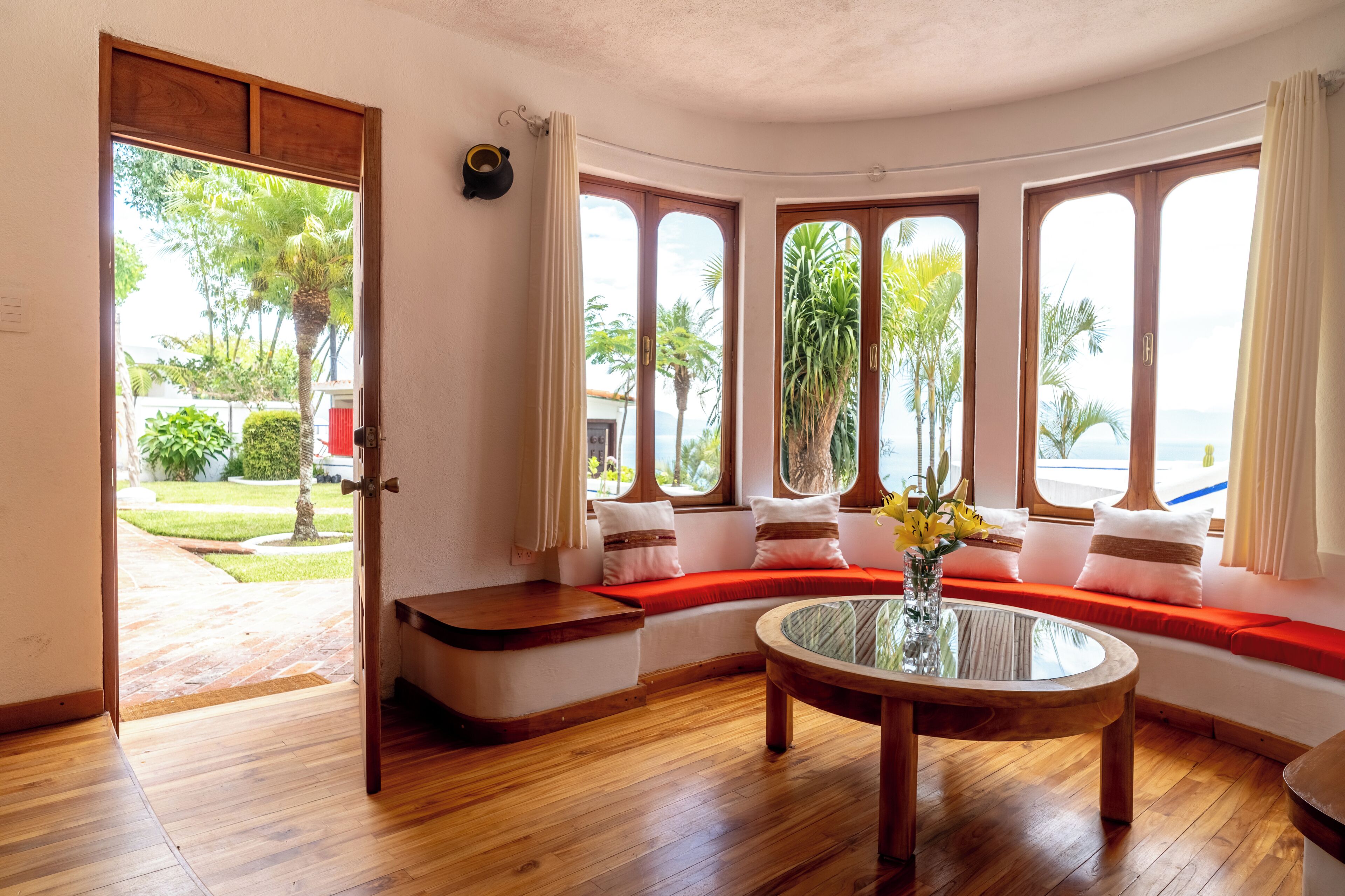 A room with a curved wooden bench, a glass coffee table, and a view of the outdoors through large windows.
