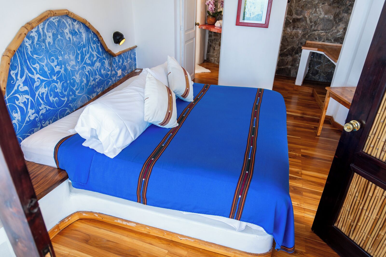 Una cama con cabecera azul y ropa de cama blanca, suelo de madera y pared de piedra al fondo.
