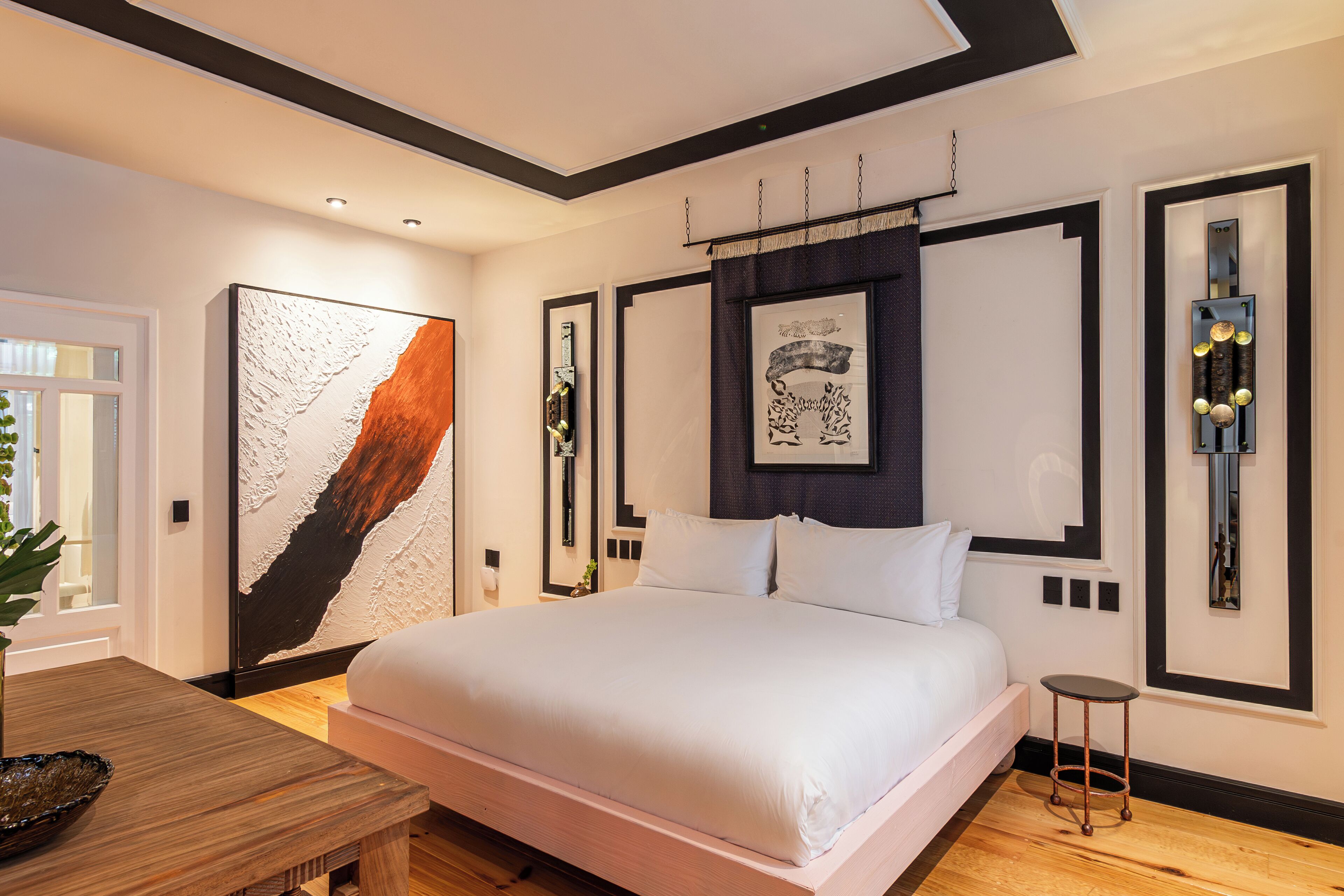 Quarto moderno com uma cama grande, mesa de cabeceira de madeira, mesinha auxiliar e obras de arte nas paredes.