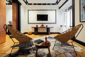 A modern living room with two wicker chairs, a small wooden table with books and a vase, a mounted TV, and a cowhide rug.