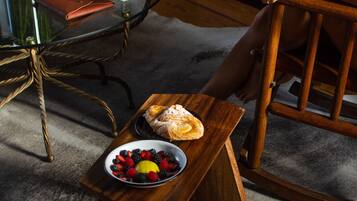 Una mesa de madera con un cuenco de fruta y un croissant, un florero de vidrio con flores, y un sombrero sobre una silla.