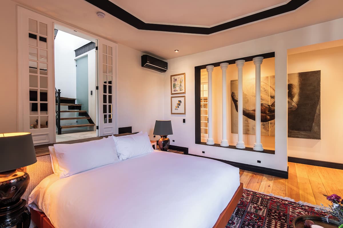 Une chambre spacieuse avec un grand lit, une table de chevet avec une lampe, un plancher en bois et une vue sur un escalier menant à une autre pièce.