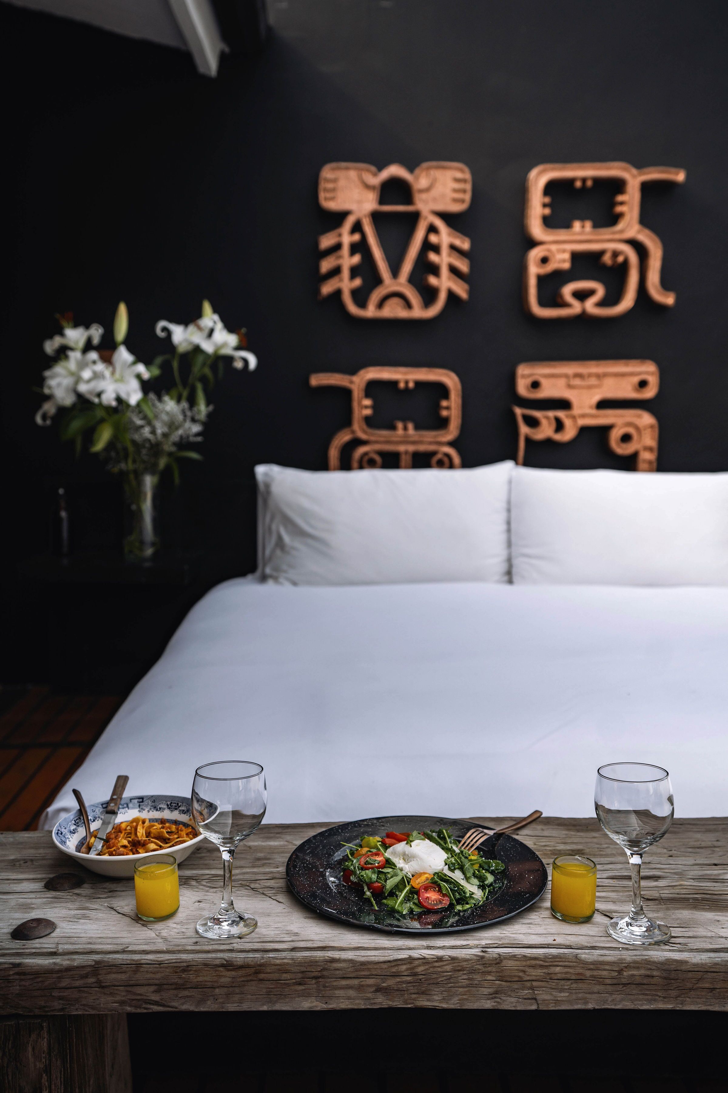 Una cama bien hecha con ropa blanca, una mesa de madera con cosas para el desayuno y una pared decorada con arte.