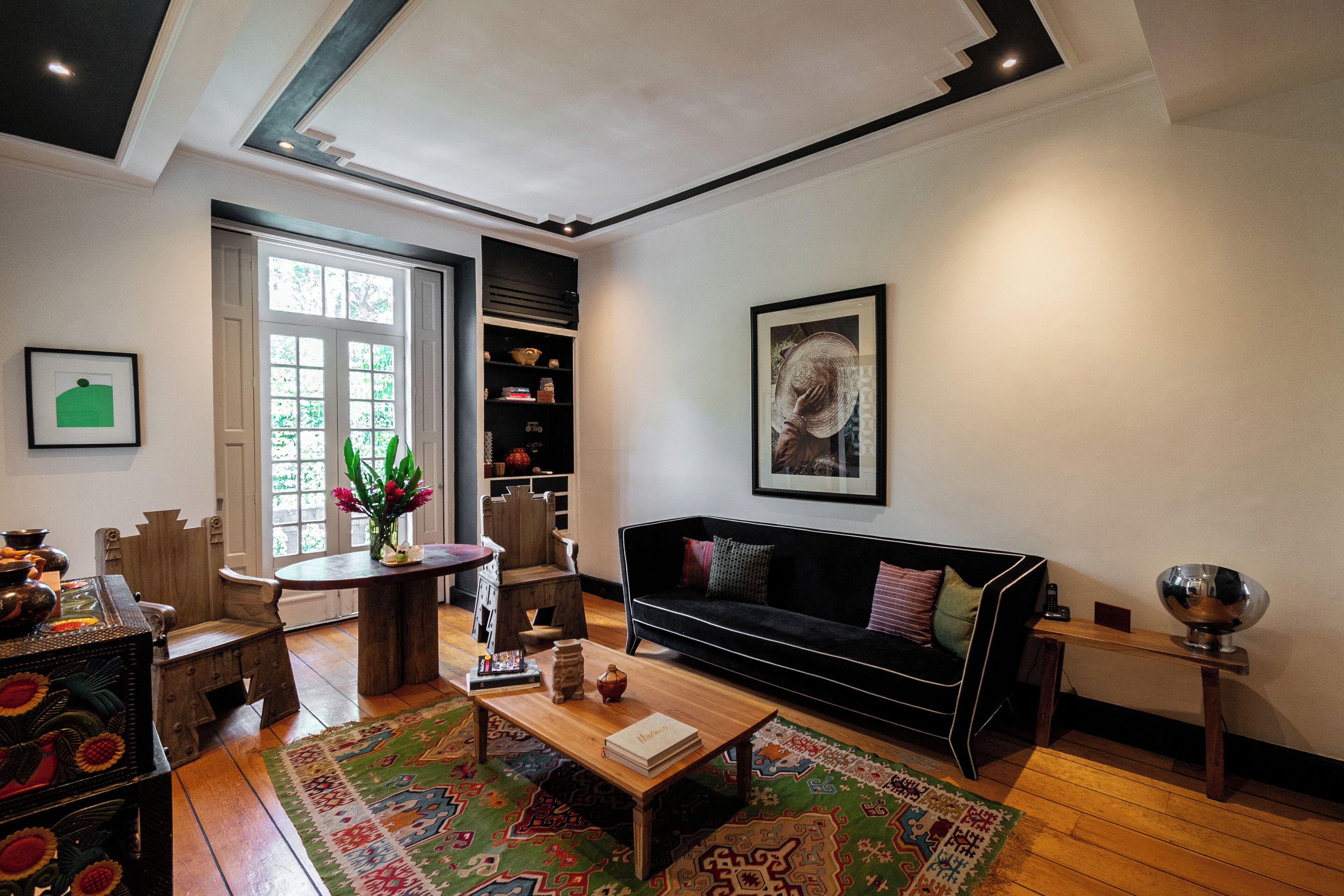Una sala de estar con un sofá negro, una mesa de centro de madera y una alfombra colorida. Se ve una estantería con varios objetos y un cuadro en la pared.