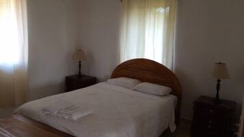 A bedroom with a wooden headboard, a single bed with white linens, two bedside tables with lamps, and a window with sheer curtains.