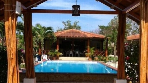 A covered pool area with a wooden pergola, a pool, and a view of a building in the background.