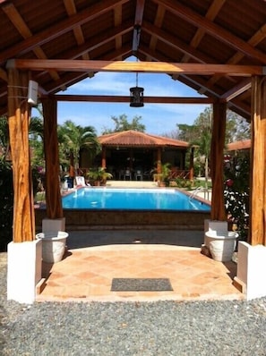 Un área de piscina cubierta con un pérgola de madera, una piscina y vista a un edificio al fondo.