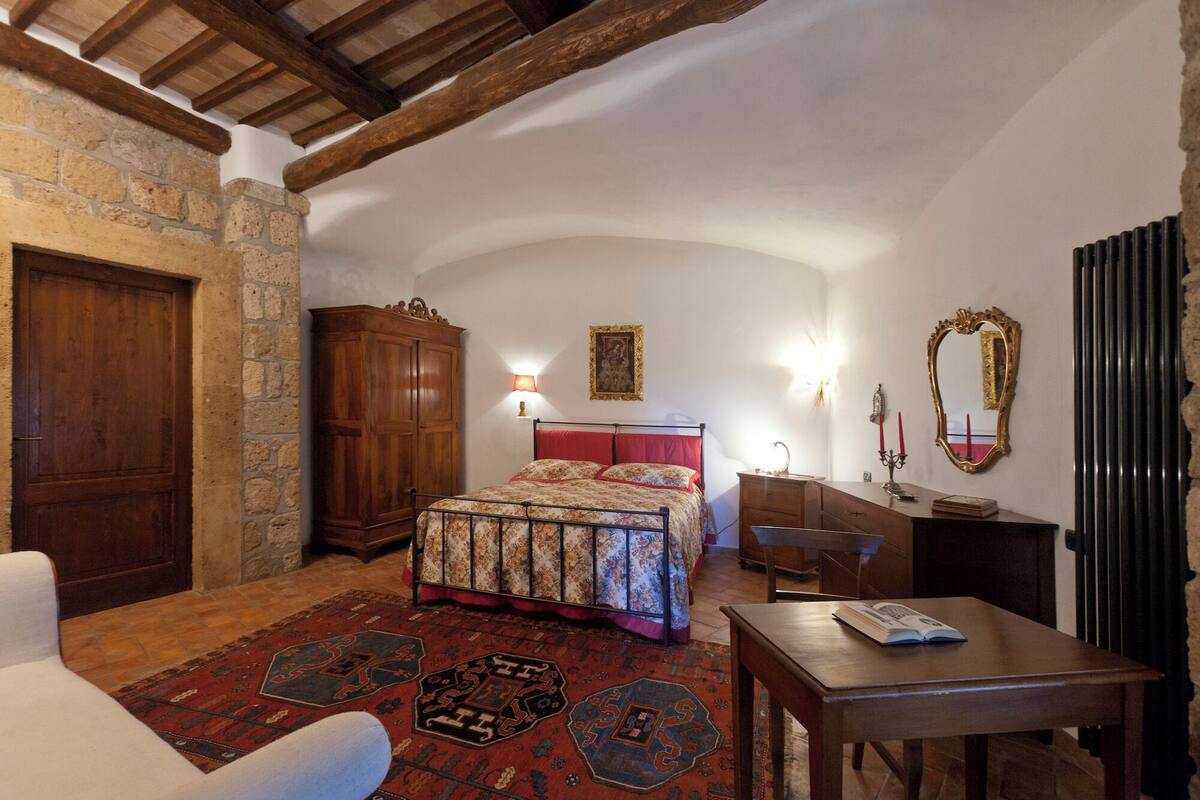 Un dormitorio con pared de piedra, armario de madera, cama con ropa de cama floral, mesita de noche, espejo y una alfombra roja.