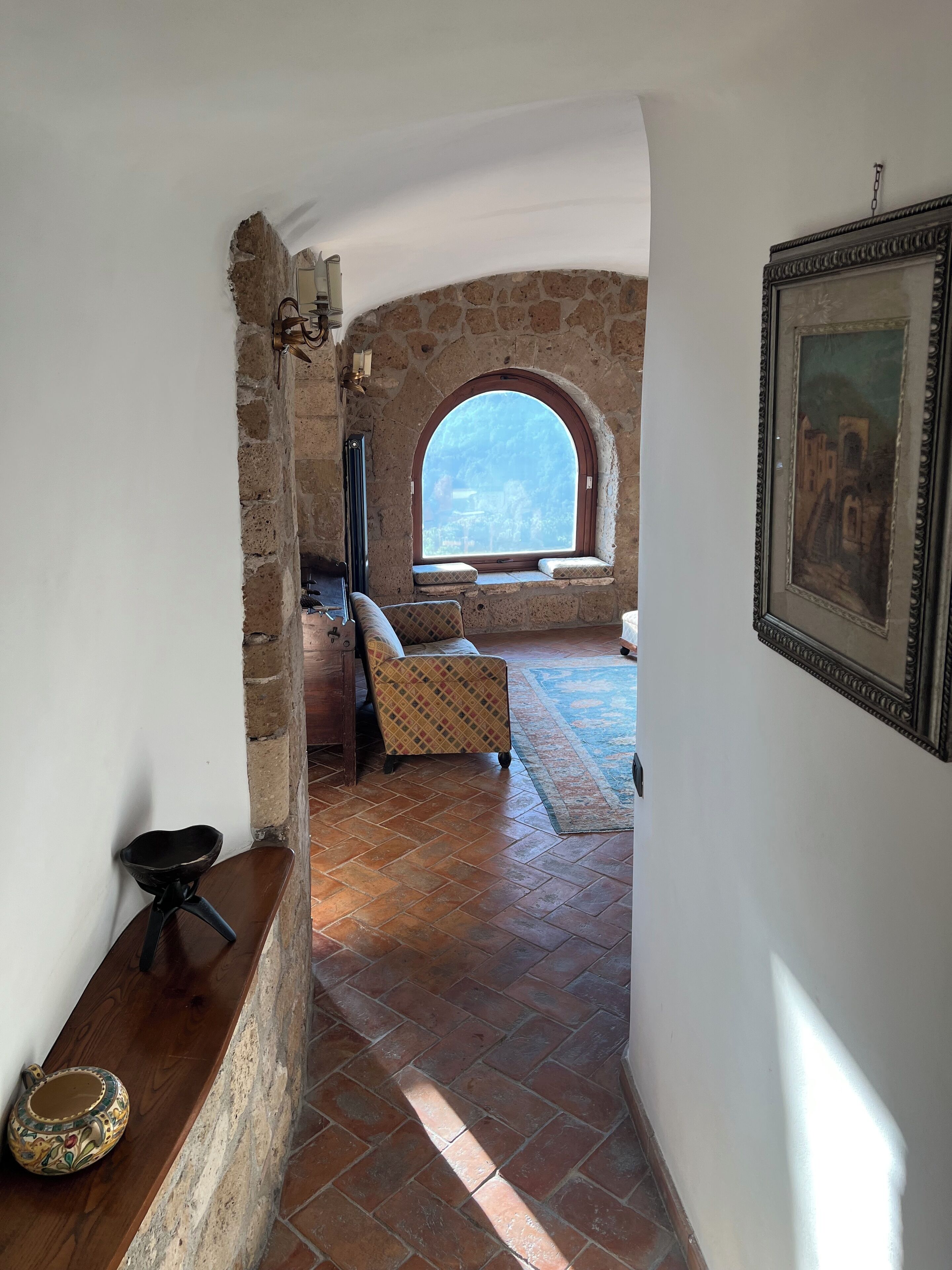 Una habitación con pared de piedra, ventana con vistas, sillón estampado y un cuadro enmarcado.