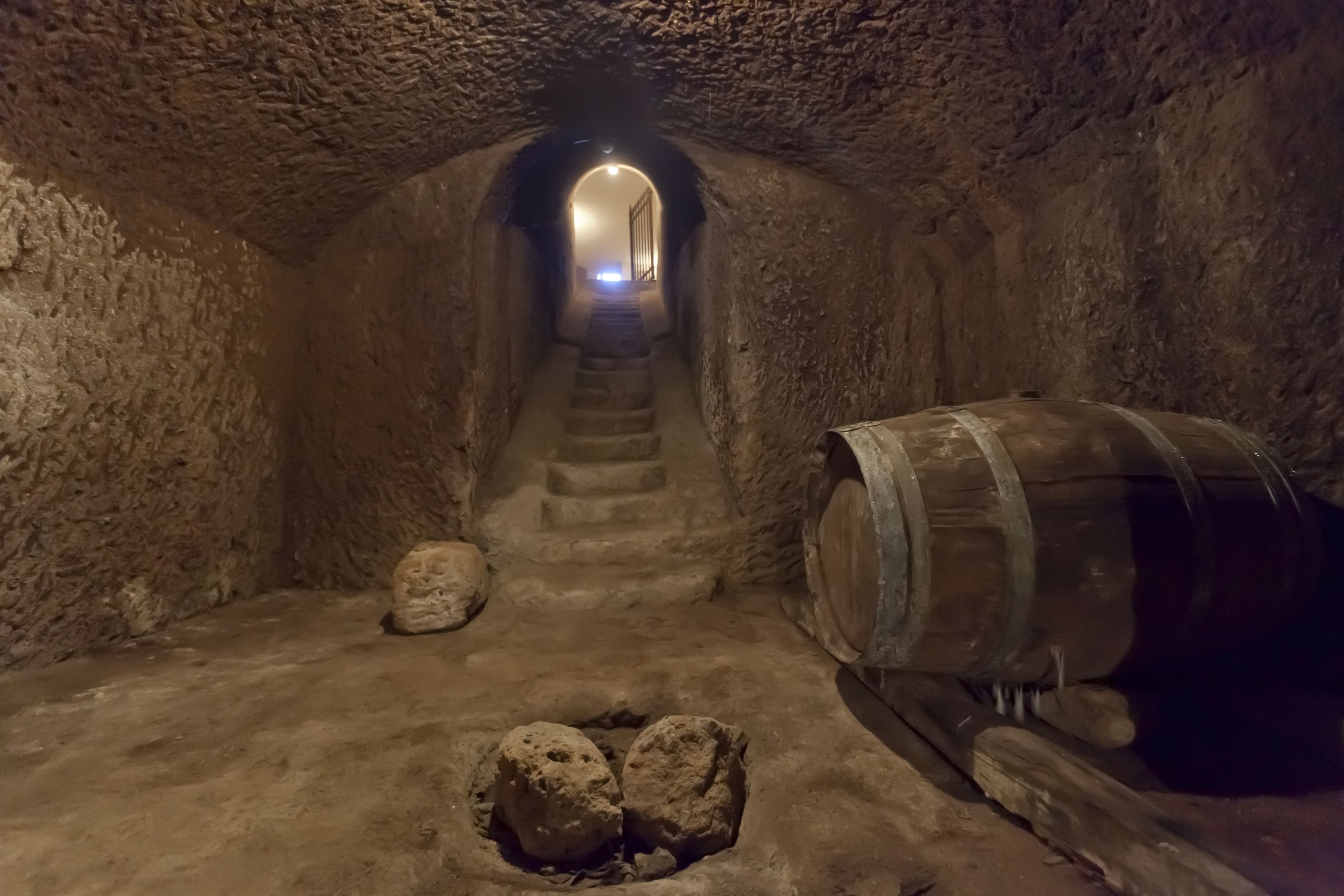 A narrow, dimly lit tunnel with a wooden barrel and two large rocks on the floor.