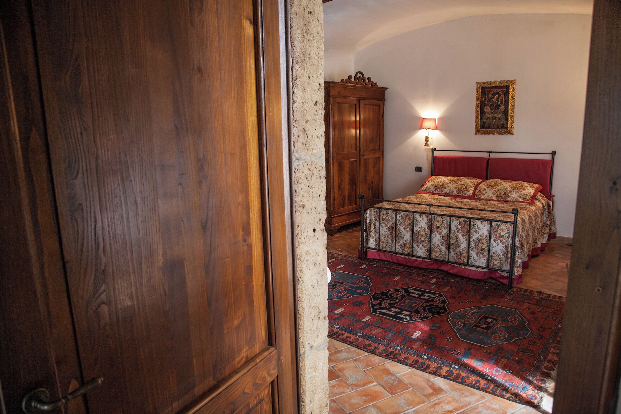 A bedroom with a wooden wardrobe, a bed with floral bedding, a wall-mounted light, and a rug with geometric patterns.
