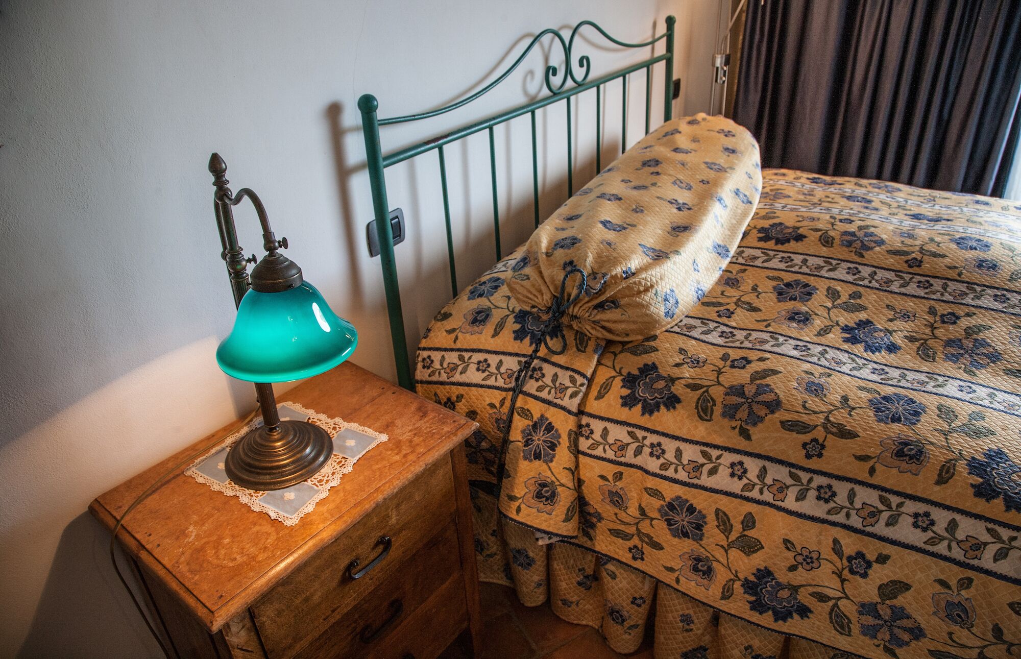 Una mesita de noche de madera con una lámpara de escritorio azul aguamarina, un copo de encaje y un cajón de madera. Una cama con edredón estampado con motivos florales y una almohada a juego, apoyada contra un cabecero de metal verde.