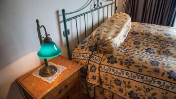 Una mesita de noche de madera con una lámpara de escritorio azul aguamarina, un copo de encaje y un cajón de madera. Una cama con edredón estampado con motivos florales y una almohada a juego, apoyada contra un cabecero de metal verde.