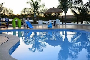 A swimming pool with a slide and a small playhouse, surrounded by palm trees and white chairs. - Enseada Flat Residence (Caldas Novas)