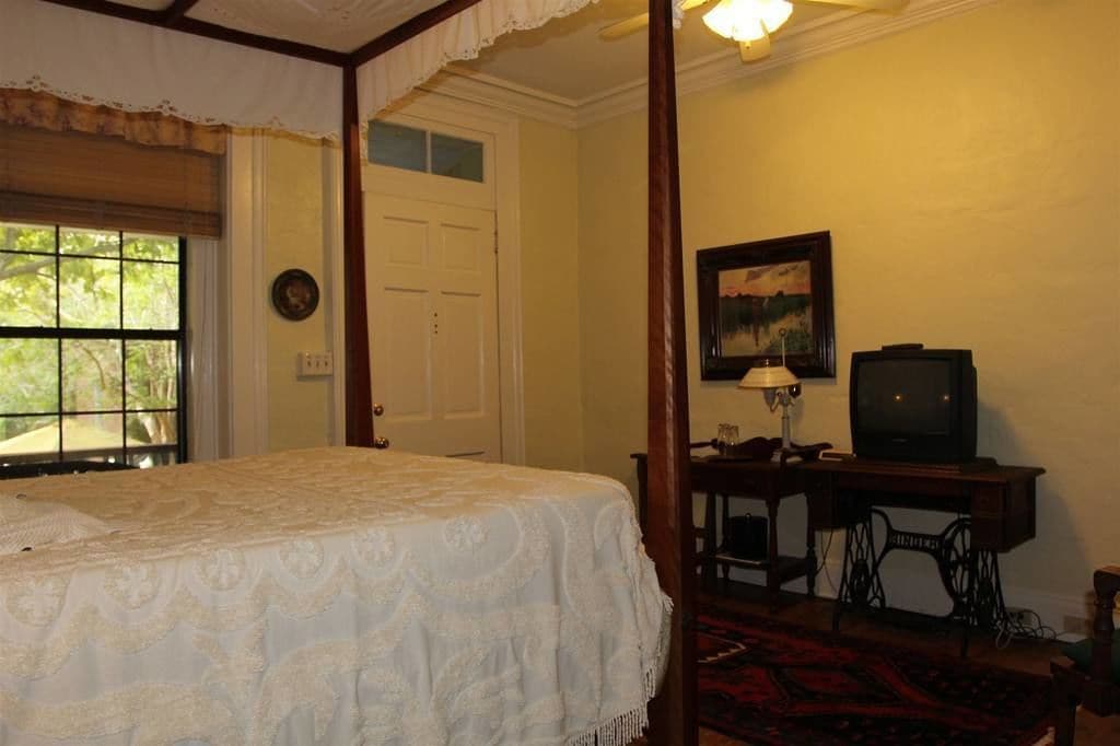 Une chambre dotée d’un lit à baldaquin, d’une machine à coudre, d’un téléviseur et d’une fenêtre donnant sur un paysage verdoyant.
