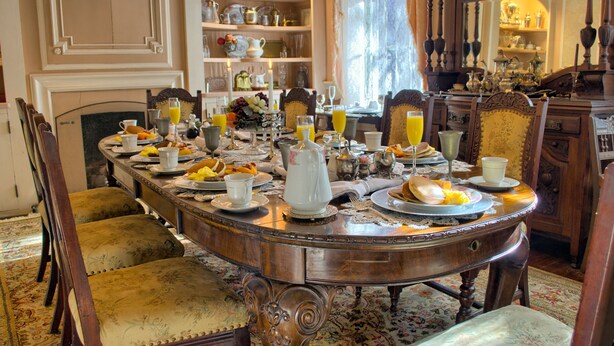 Une salle à manger élégante, avec une longue table dressée pour le petit-déjeuner, entourée de chaises richement décorées et agrémentée d’un miroir ornemental.