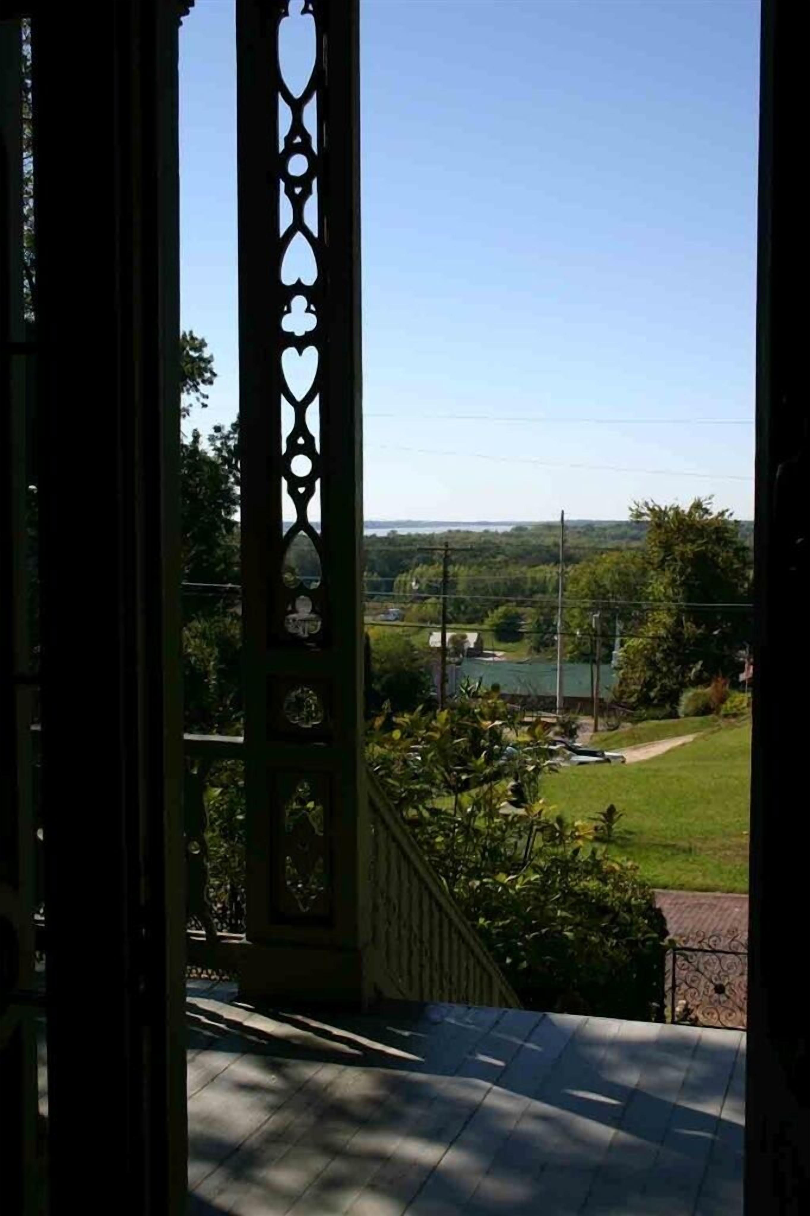 A view from a balcony with a railing featuring decorative cutouts, overlooking a green landscape with a clear sky.