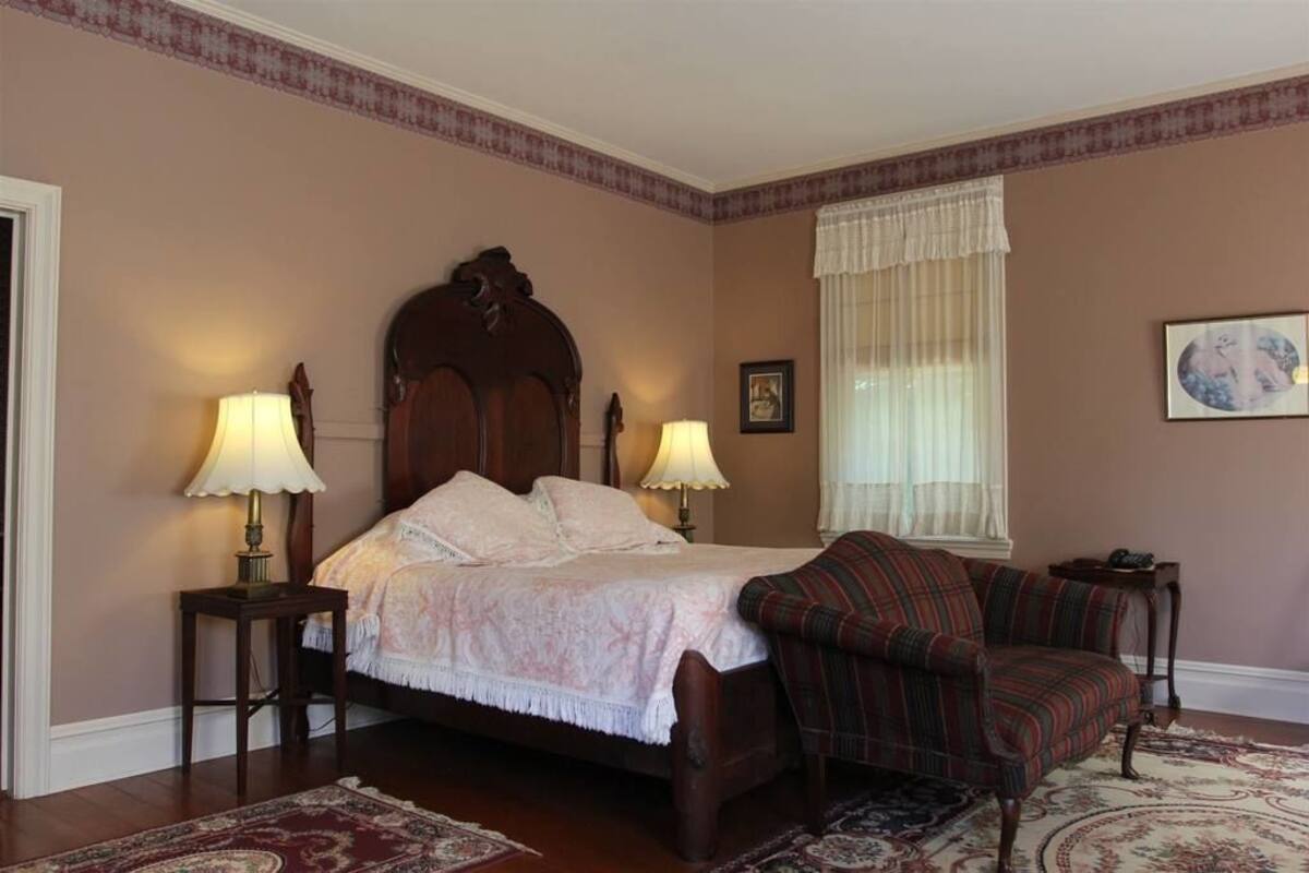Un dormitorio con una cama de madera, dos lámparas de noche, un sillón a cuadros, una ventana con cortinas de encaje y un cuadro en la pared.