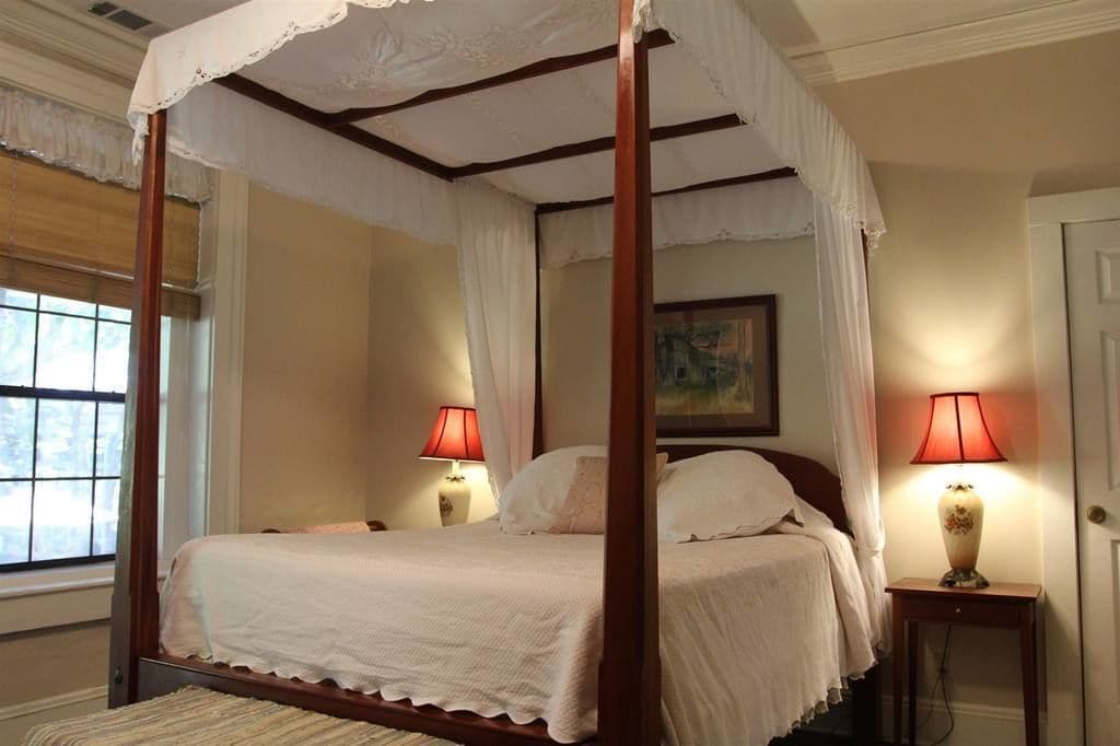 A four-poster canopy bed with two bedside tables, each with a lamp, in a room with a window and a painting on the wall.