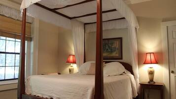 A four-poster canopy bed with two bedside tables, each with a lamp, in a room with a window and a painting on the wall.