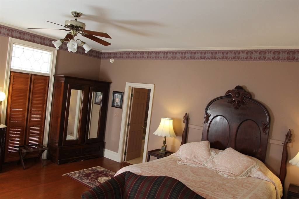 A bedroom with a bed, a ceiling fan, a wooden wardrobe, a mirror, and a lamp.