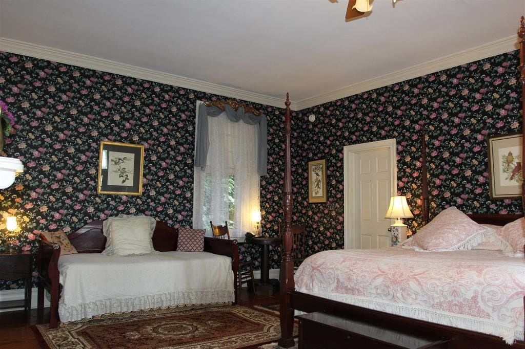 A traditional bedroom with floral wallpaper, two beds, a four-poster bed, and a nightstand with a lamp.