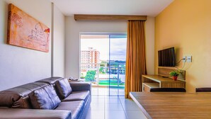 A modern living room with a leather sofa, a dining table, a TV, and a large window with a view of buildings and greenery.