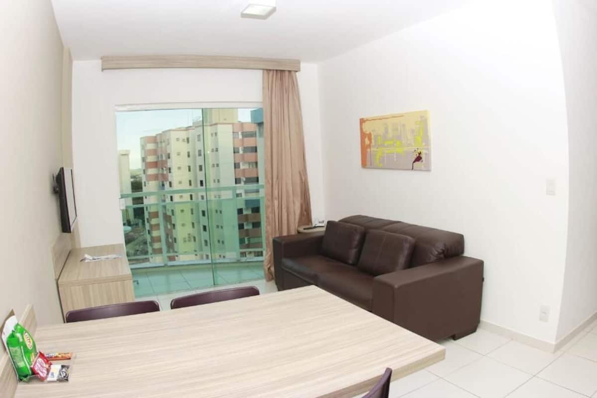 A modern living room with a brown leather sofa, a dining table, and a large window overlooking a cityscape.