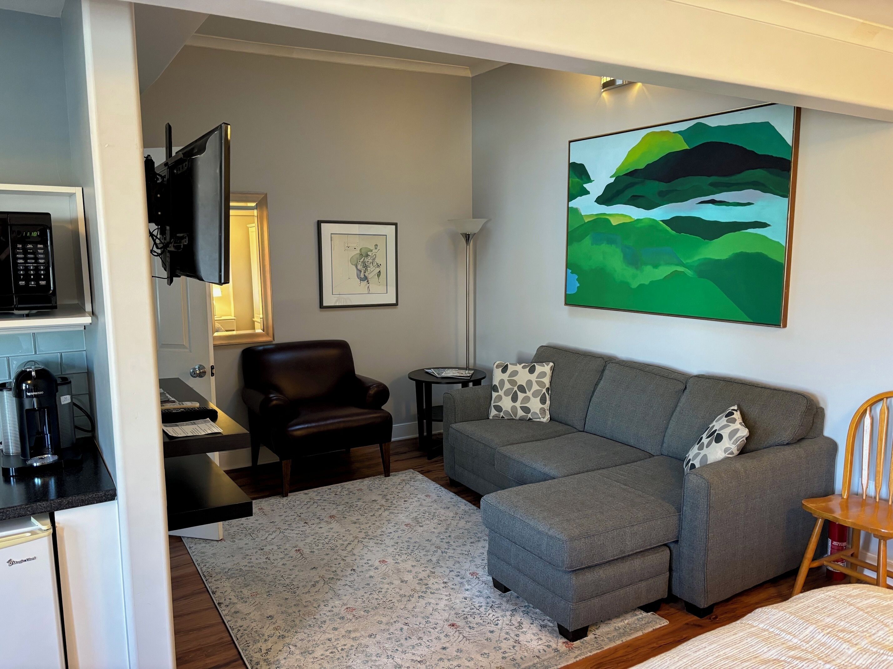 A modern living room with a grey sectional sofa, a black leather chair, a small side table, a coffee machine, and a painting on the wall.