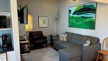 A modern living room with a grey sectional sofa, a black leather chair, a small side table, a coffee machine, and a painting on the wall.