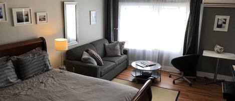A hotel room with a bed, a sofa, a desk, and a chair. There are pictures on the walls and a window with curtains.