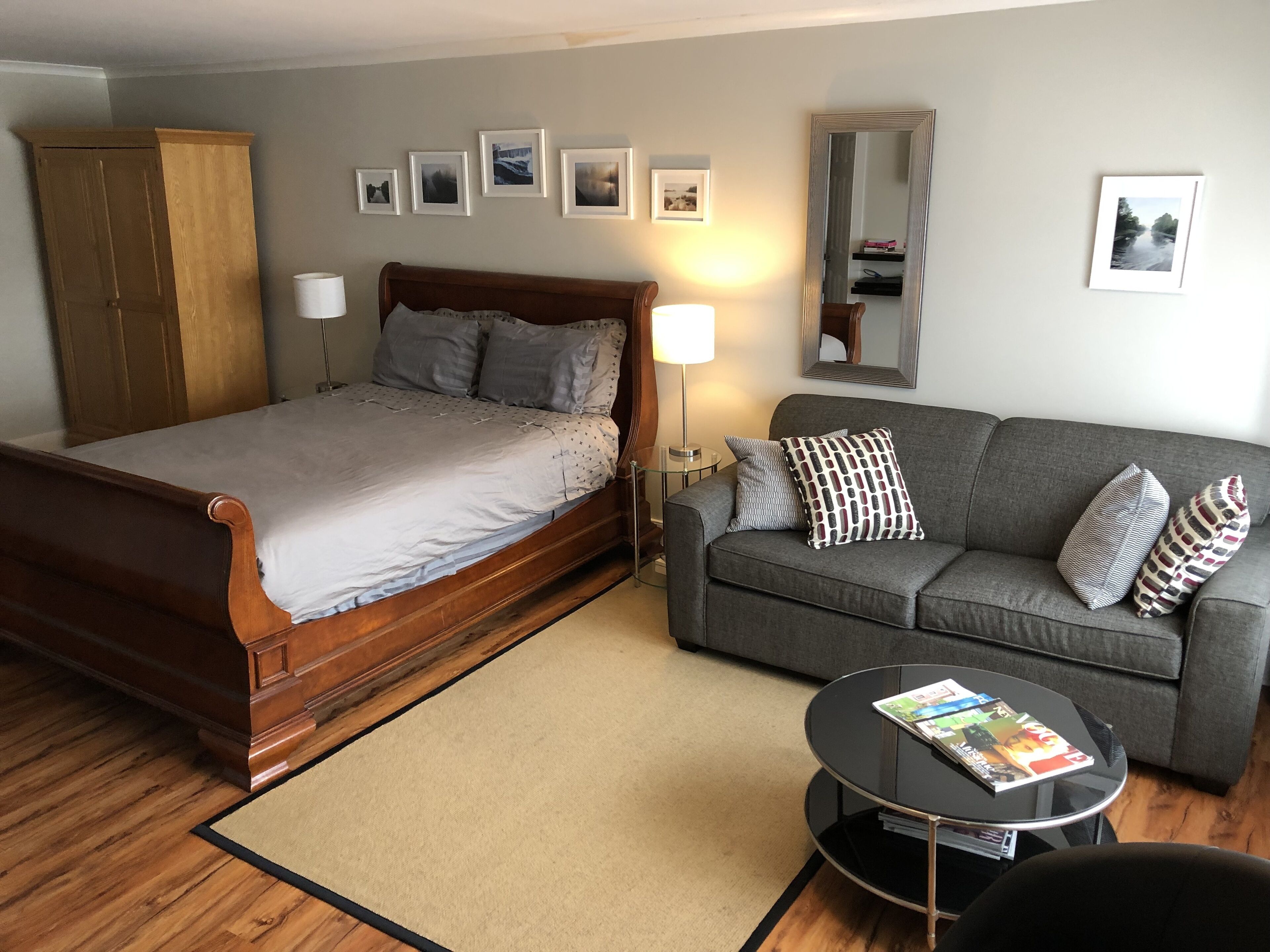 A bedroom with a wooden bed, a grey sofa, a small round table with magazines, and a mirror on the wall.
