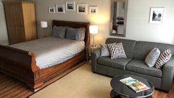 A bedroom with a wooden bed, a grey sofa, a small round table with magazines, and a mirror on the wall.