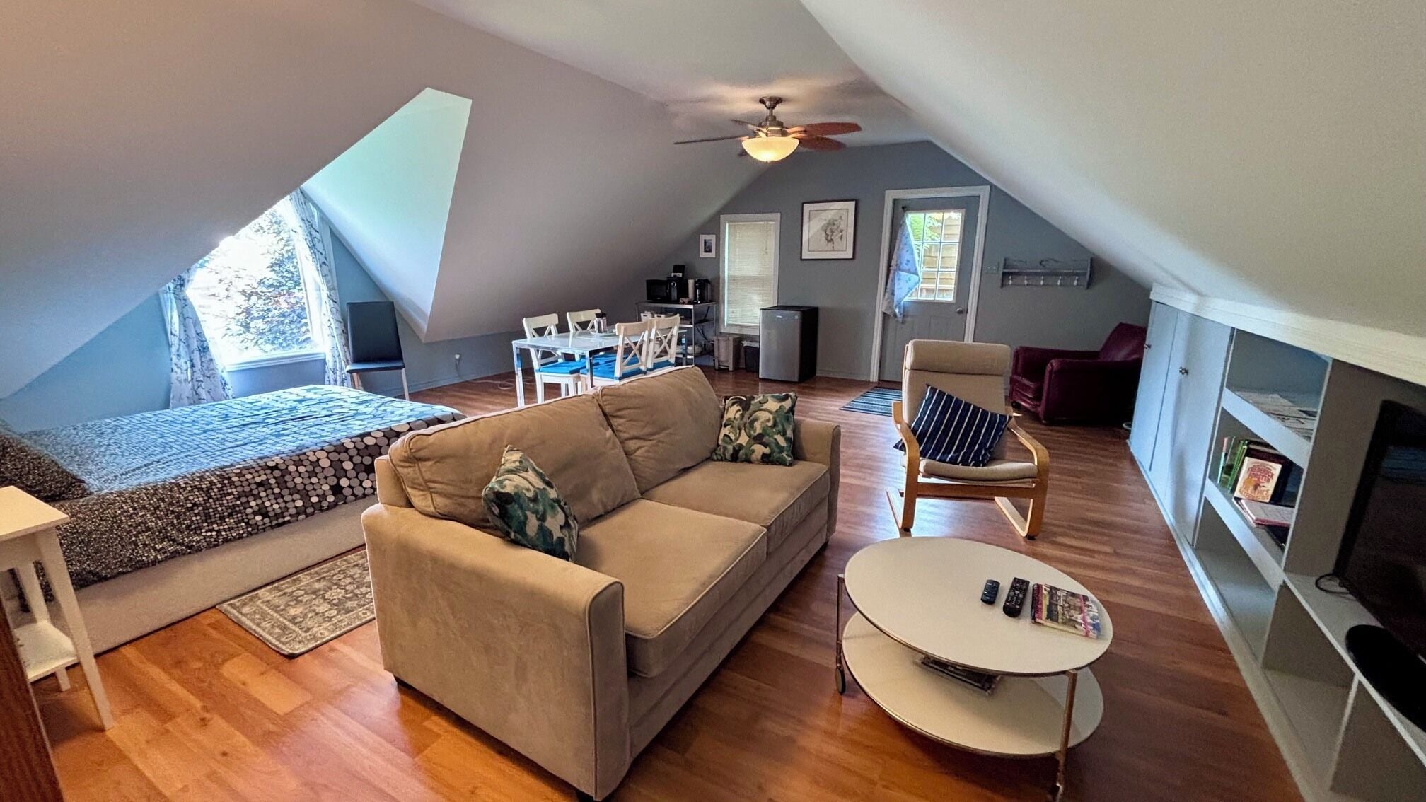 A spacious loft with a high ceiling, a cozy seating area with a sofa and armchair, a dining table, and a bookshelf.