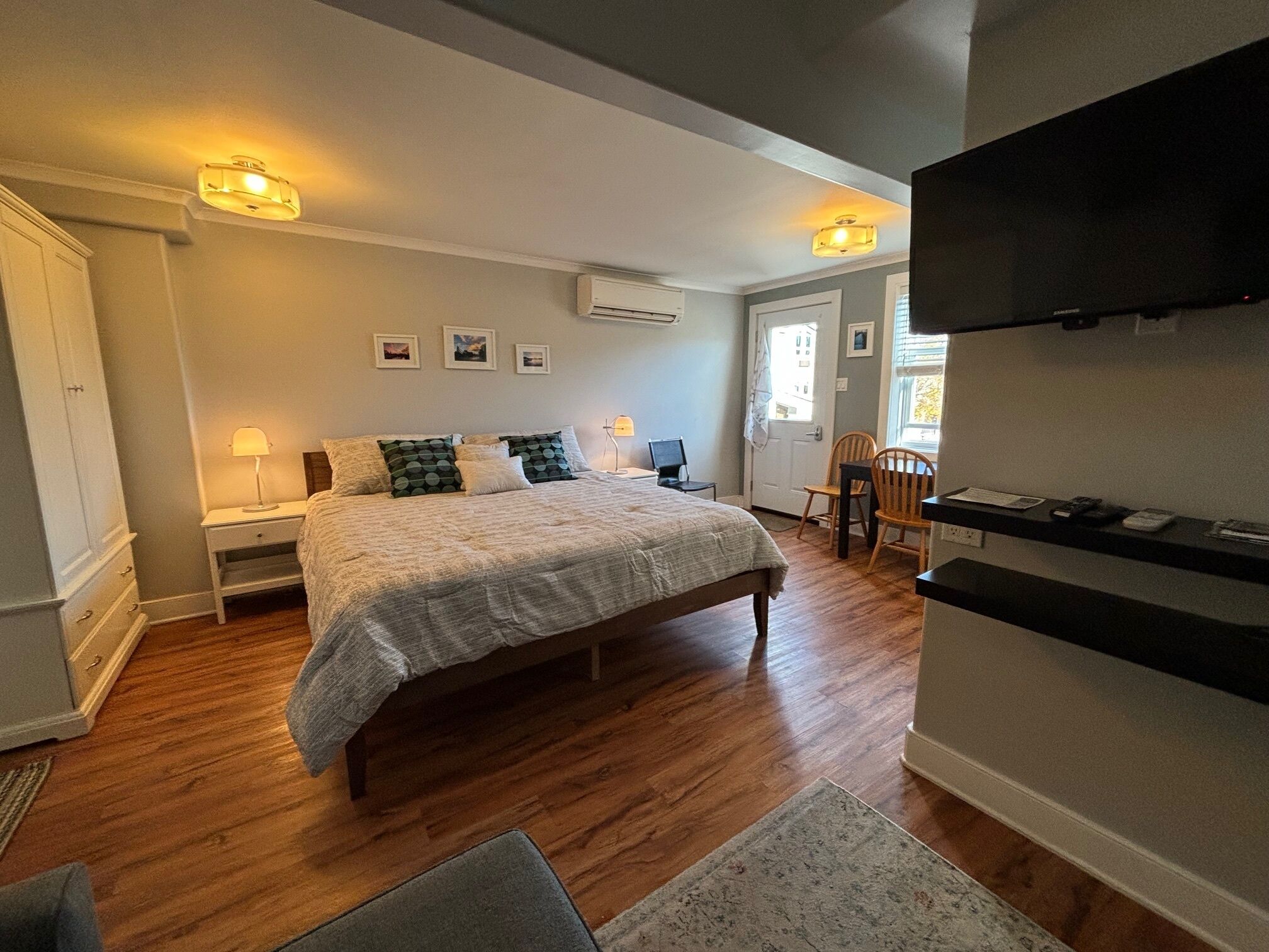 Une chambre à coucher comprenant un lit, des tables de chevet, un bureau et une chaise. On y trouve également des lampes fixées au mur, un téléviseur à écran plat et une porte donnant sur une autre pièce.