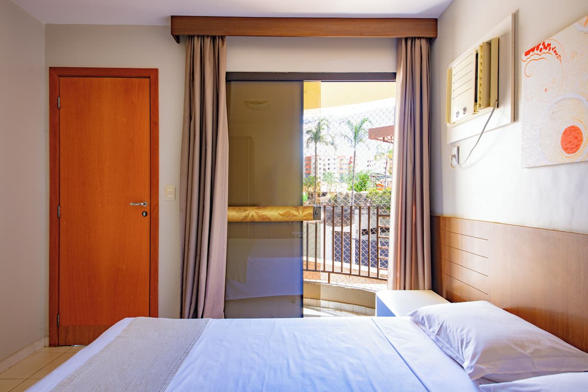 A hotel room with a wooden door, a balcony with a view of palm trees and buildings, and a bed with white linens.