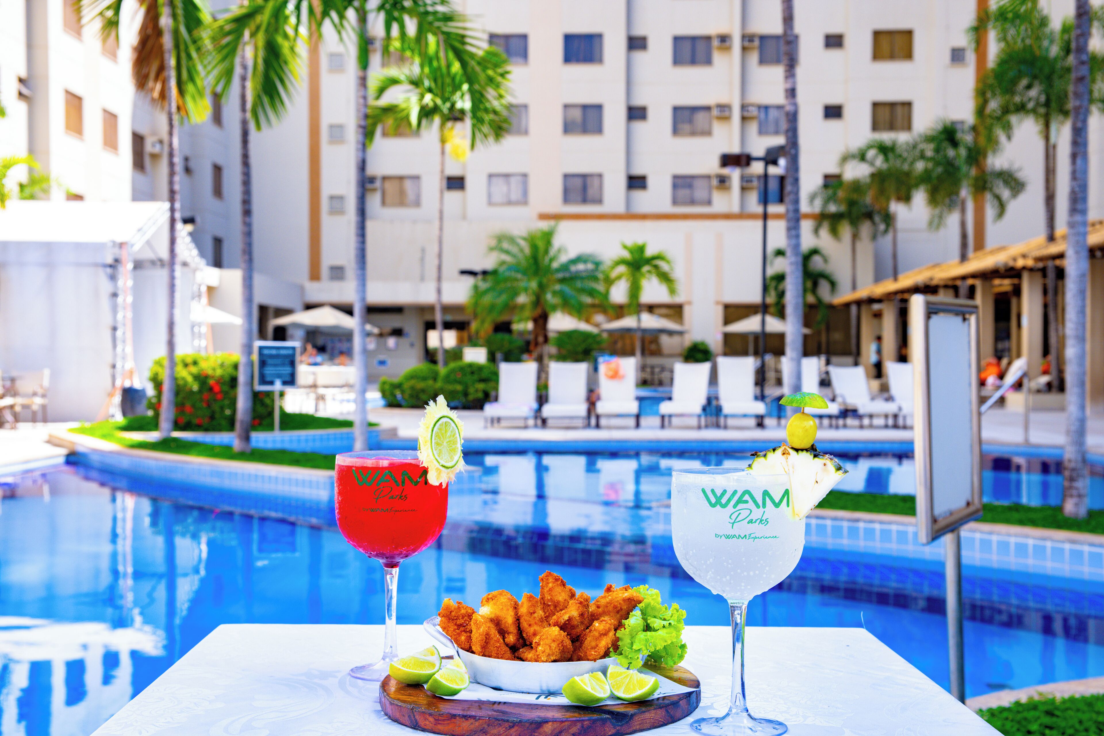 Mesa junto a la piscina con dos bebidas, un bol de comida frita y vistas a un edificio de hotel con palmeras.