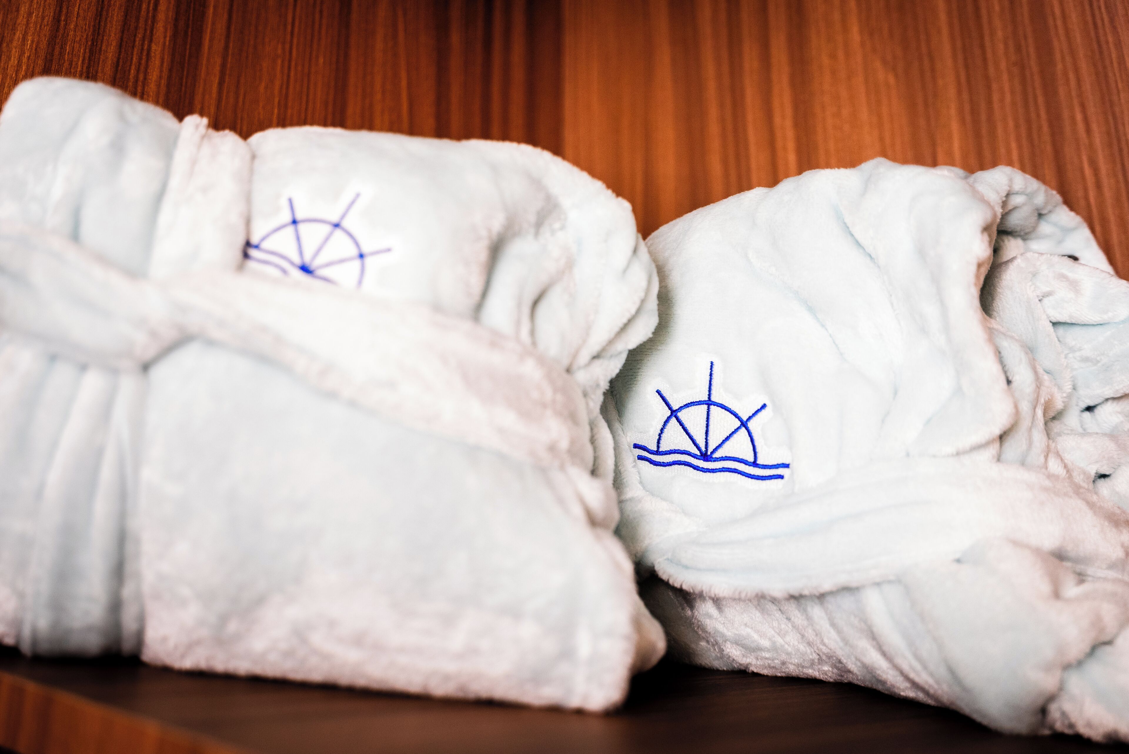 Two white bathrobes with blue embroidery on a wooden shelf.