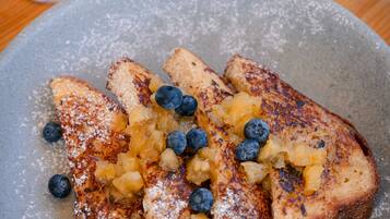 A plate of French toast with blueberries and powdered sugar, a bowl of fruit, and a glass of orange juice on a wooden table.