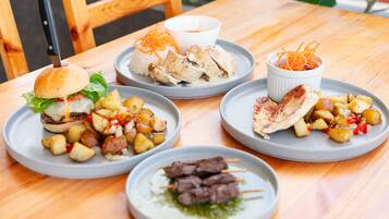 A wooden table with various dishes including a burger, chicken, skewers, and potato dishes.