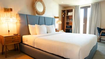 A neatly made bed with a blue headboard, a bedside table with a lamp, a mirror on the wall, and a wardrobe with hanging clothes.