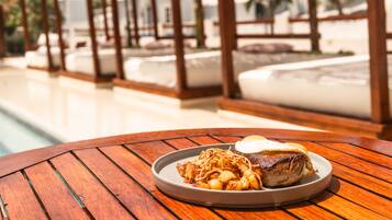 Una mesa de madera con un plato de comida, una piscina y un pabellón al fondo.